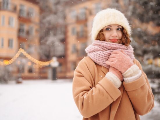 As it snows, a woman clutches her knit scarf to her chest, her wool coat, jacket, and gloves keeping her warm.