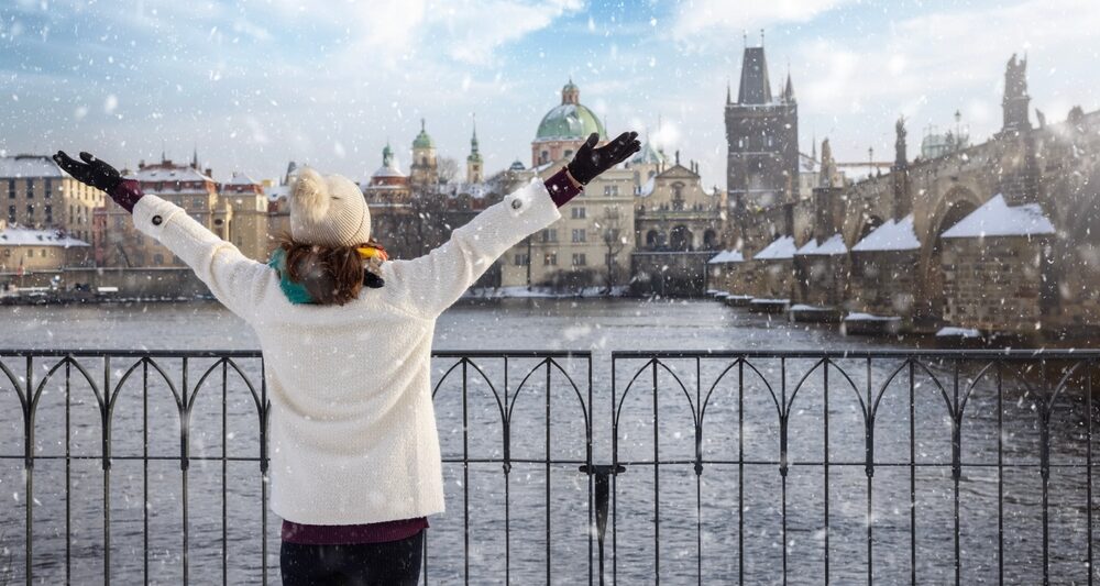 A woman looks across a river as it snows, and she knows what to wear in Europe in winter, as she is prepared with her jacket, beanie, gloves and hat.