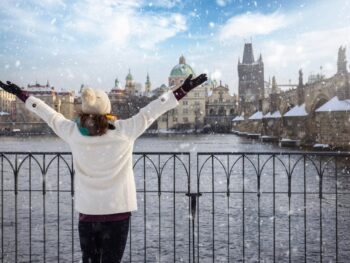 A woman looks across a river as it snows, and she knows what to wear in Europe in winter, as she is prepared with her jacket, beanie, gloves and hat.
