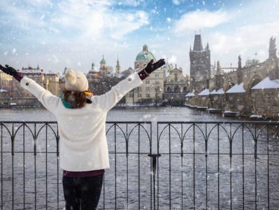 A woman looks across a river as it snows, and she knows what to wear in Europe in winter, as she is prepared with her jacket, beanie, gloves and hat.
