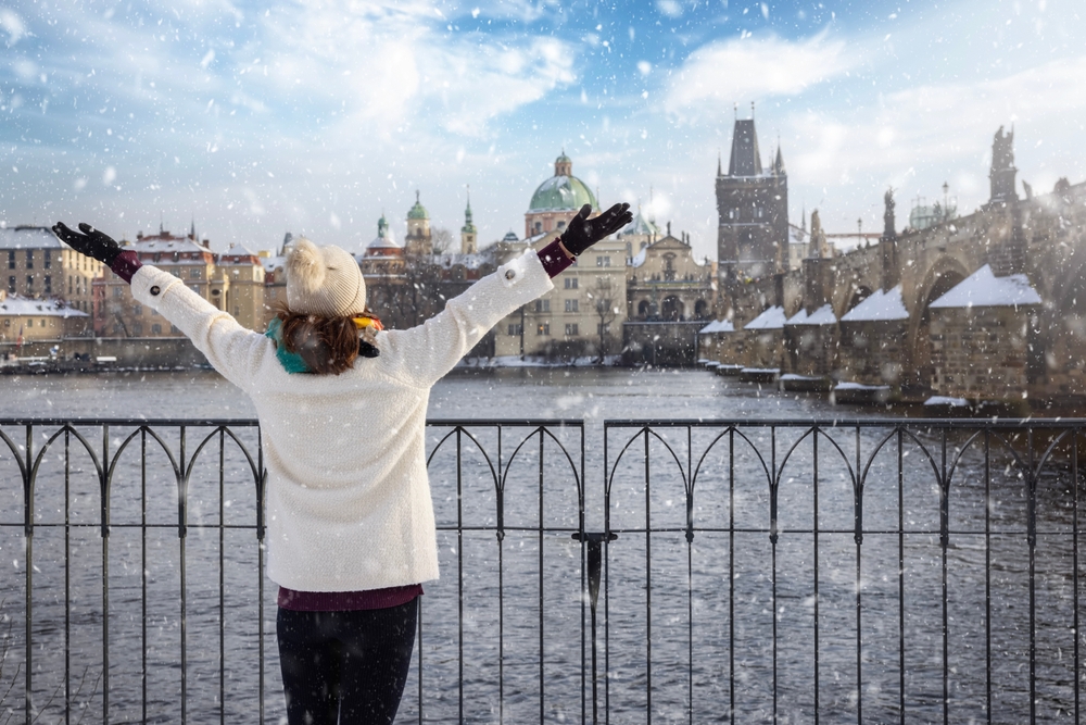 A woman looks across a river as it snows, and she knows what to wear in Europe in winter, as she is prepared with her jacket, beanie, gloves and hat.