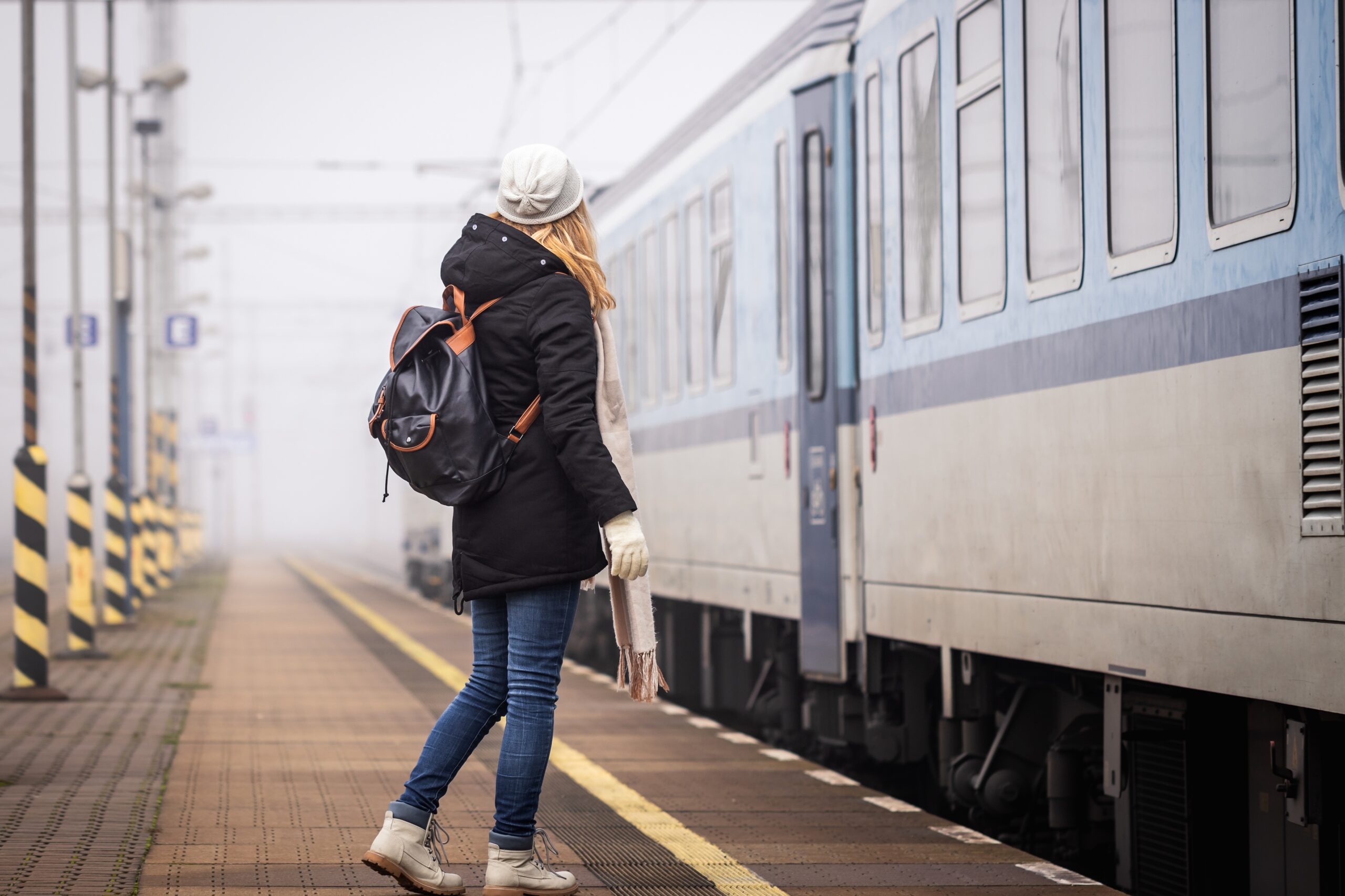 A woman waiting to board public transportation knows she knows what to wear in Europe in winter in her boots, jeans, long parka, scarf, beanie and gloves.