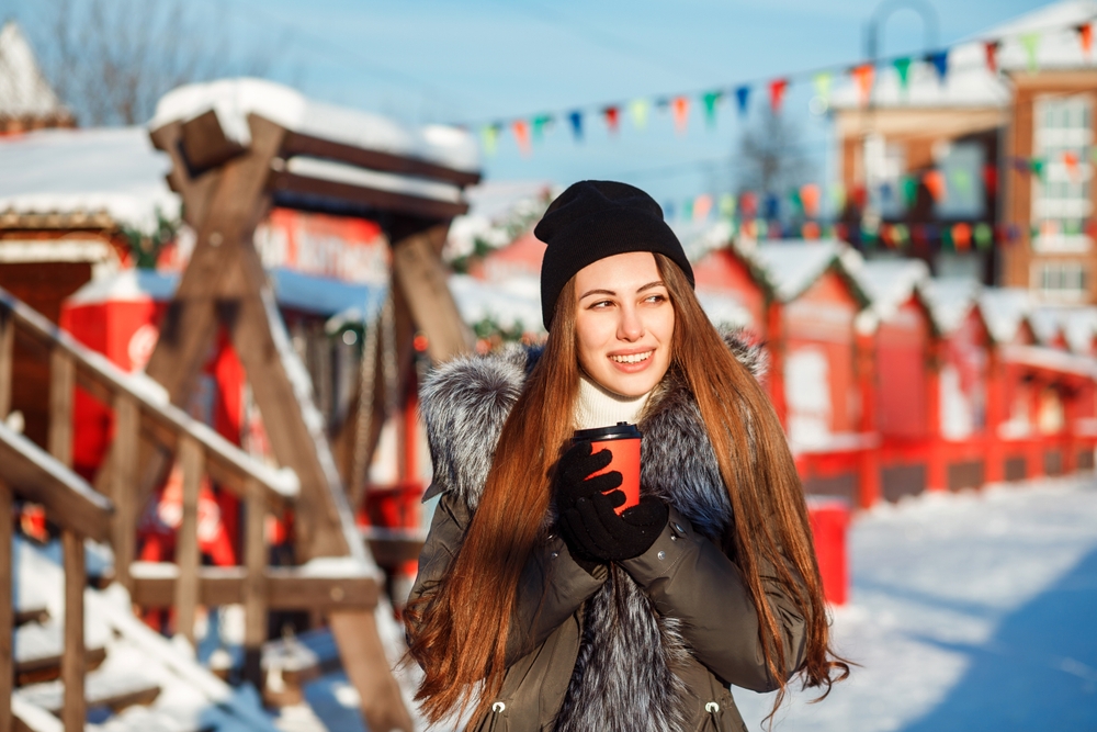A woman holds a cup of coffee beyond holiday market stalls while wearing a beanie, a fur lined parka, and gloves.