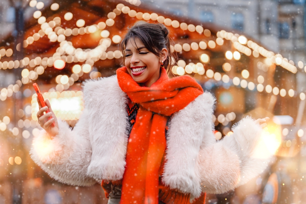 As it snows behind her, a woman smiles at her phone, knowing What to Wear in Italy in December with her cropped sherpa jacket and red scarf.