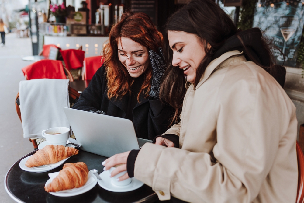 Two friends huddle outside a cafe in the cold, knowing what to wear in Europe in winter in their parks, wearing gloves, while drinking coffee and eating croissants.