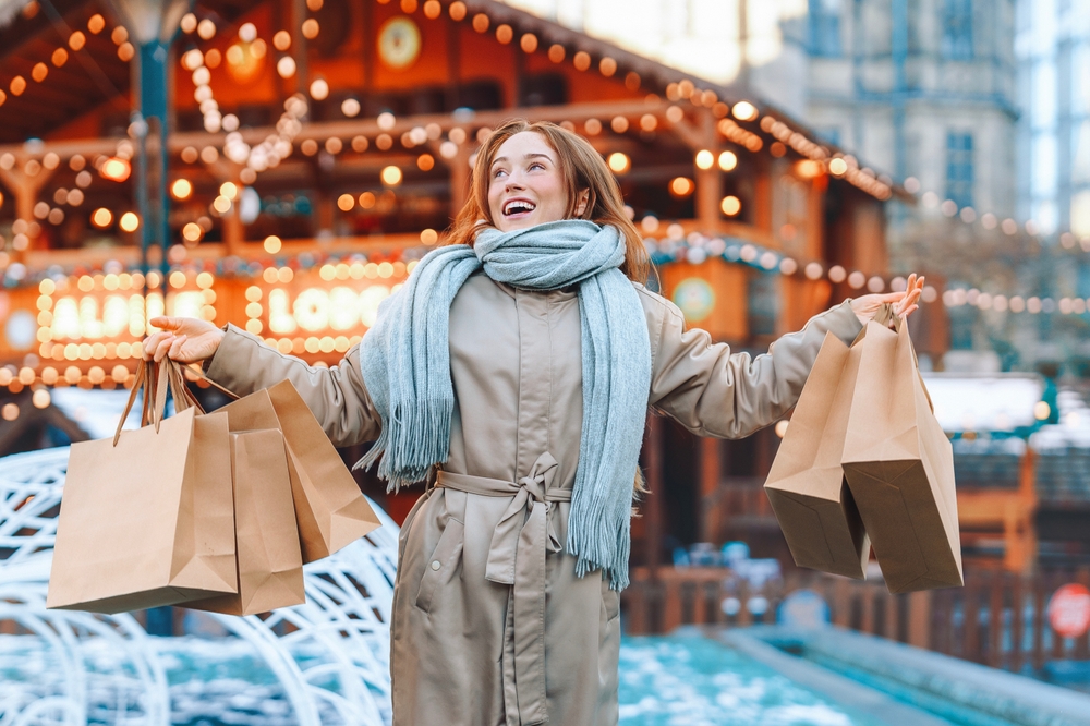 A woman gallops away from a brightly light building in her thick, waterproof trench coat and blue scarf, her hands full of shopping bags.