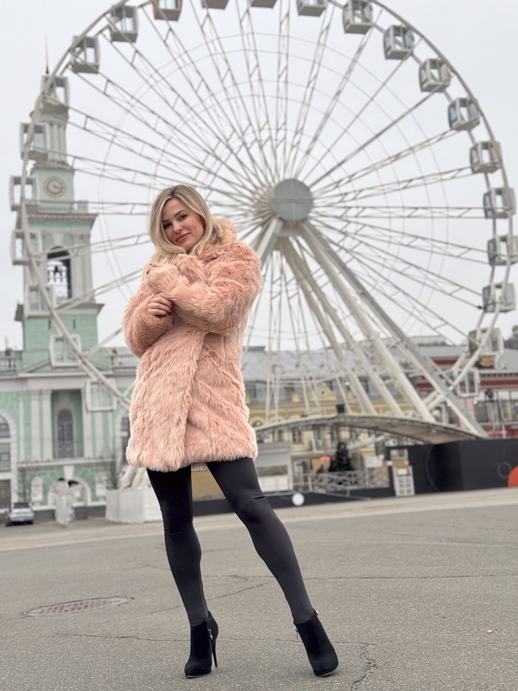 A woman stands in front of a giant ferris wheel in a sherpa trench coat, leggings and boots.