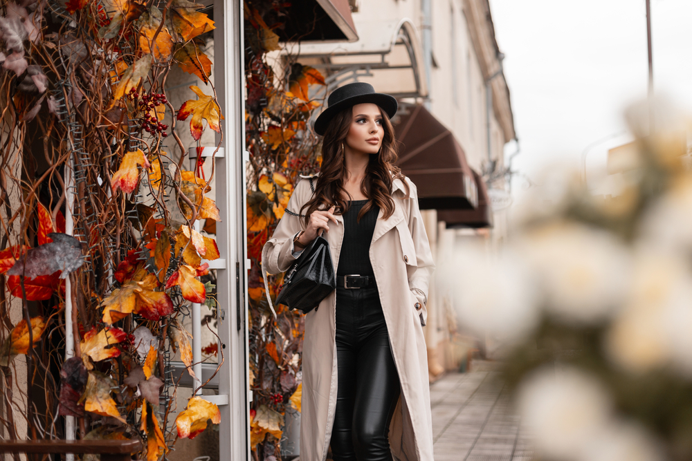 A woman walks down a street where fall leaves turn and decorate buildings. She wears leather pants and a trench coat with a hat and structured purse.