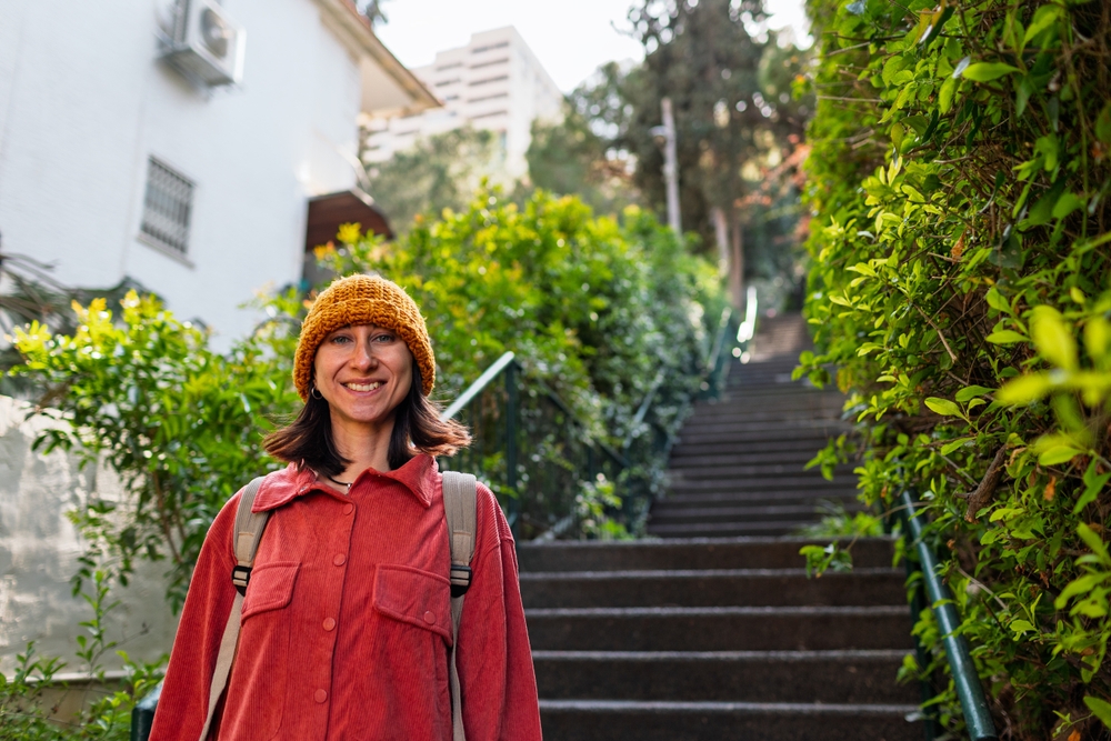 At the bottom of stairs, beyond greenery, a woman smiles in her orange beanie and button down fleece.