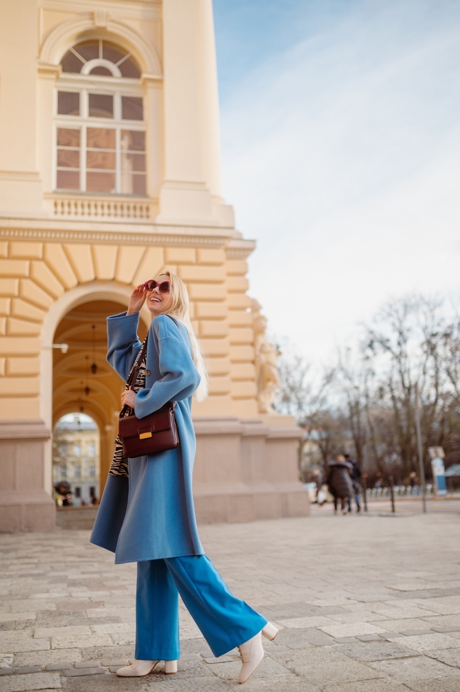 A woman knows What to Wear in Italy in fall as she smiles over her shoulder while wearing a long blue coat with matching pants.