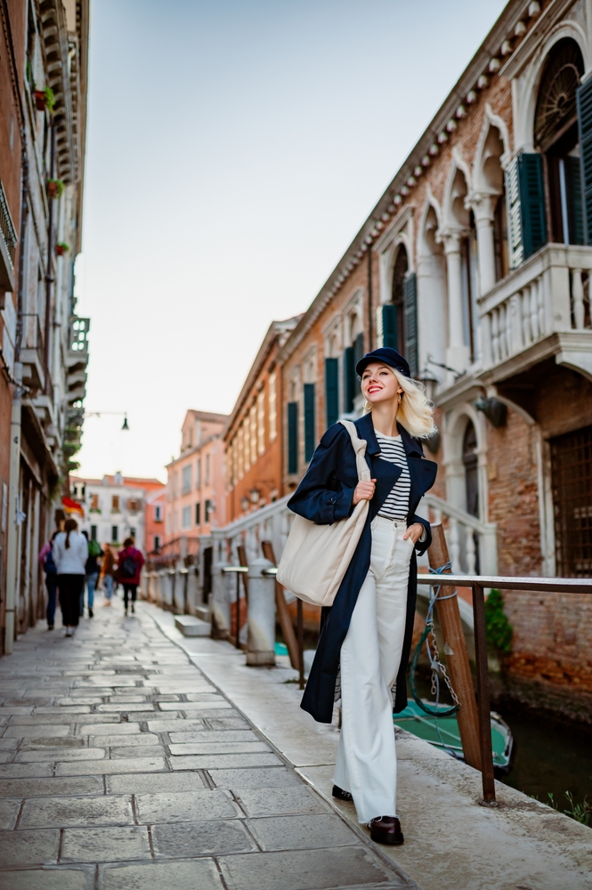 A woman walks down a side street and canal in her blue trench, white jeans, striped shirt and hat: she knows What to Wear in Italy in Fall.