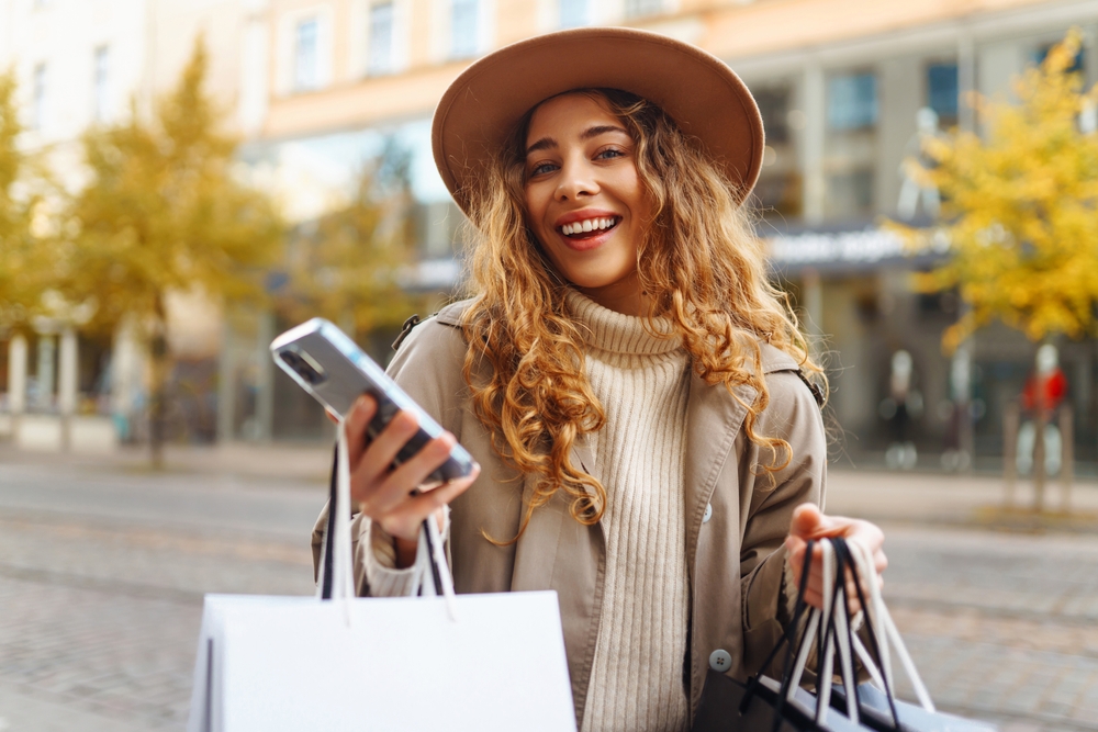 A young girl with curls smiles at the camera with her turtleneck, jacket and hat all in monochrome colors, her hands full of shopping bags.