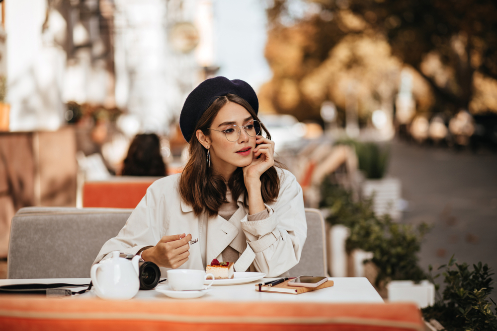 A woman sits outside at a cafe enjoying a treat and a coffee while wearing a hat, trench and glasses.