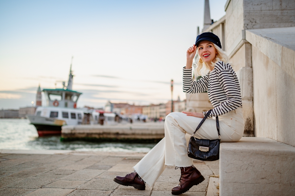 A woman sits on the edge of a step on the coast of Italy in jeans, boots, a hat and a striped shirt.