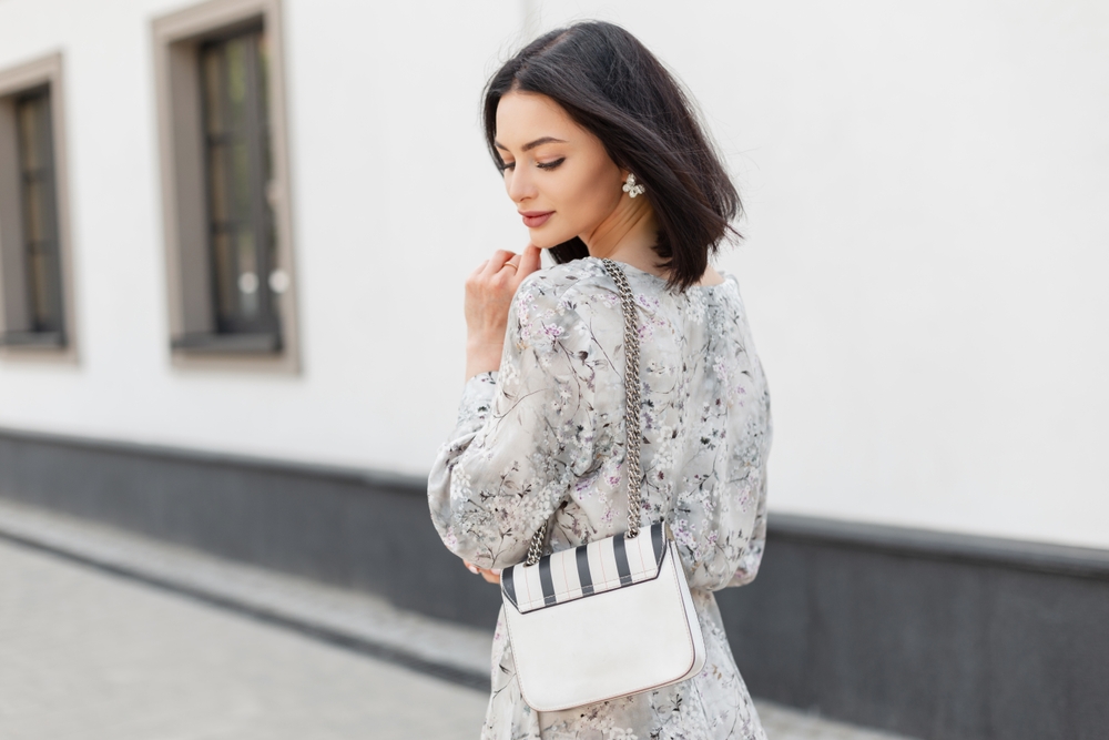 A woman glances over her shoulder while wearing a floral dress and carrying a structured purse over her shoulder.
