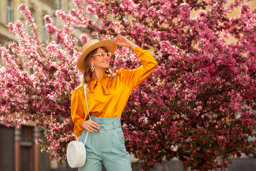 This woman poses in front of floral blooms, enjoying spring and knowing What to wear in Italy in spring in her silk yellow top and high waisted pants.