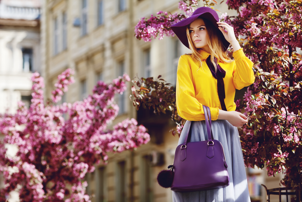 This woman knows What to wear in Italy in spring as she poses in front of flower blooms, her hat and purse matching the purple flowers behind her.