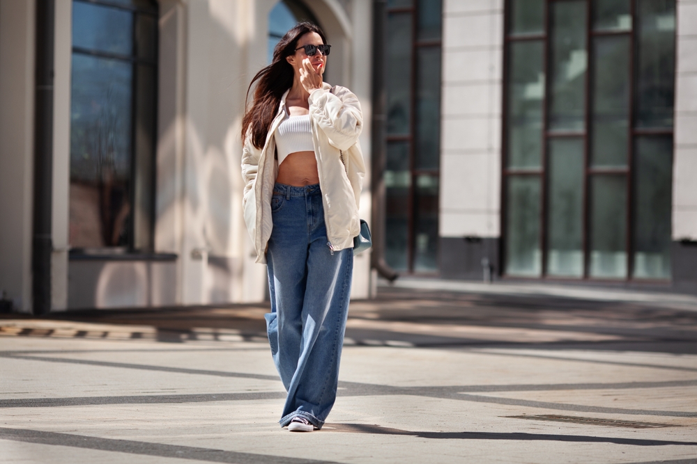 A woman walks down the street, looking casual yet stylish, in her high waisted jeans, parka, cropped top and sneakers.