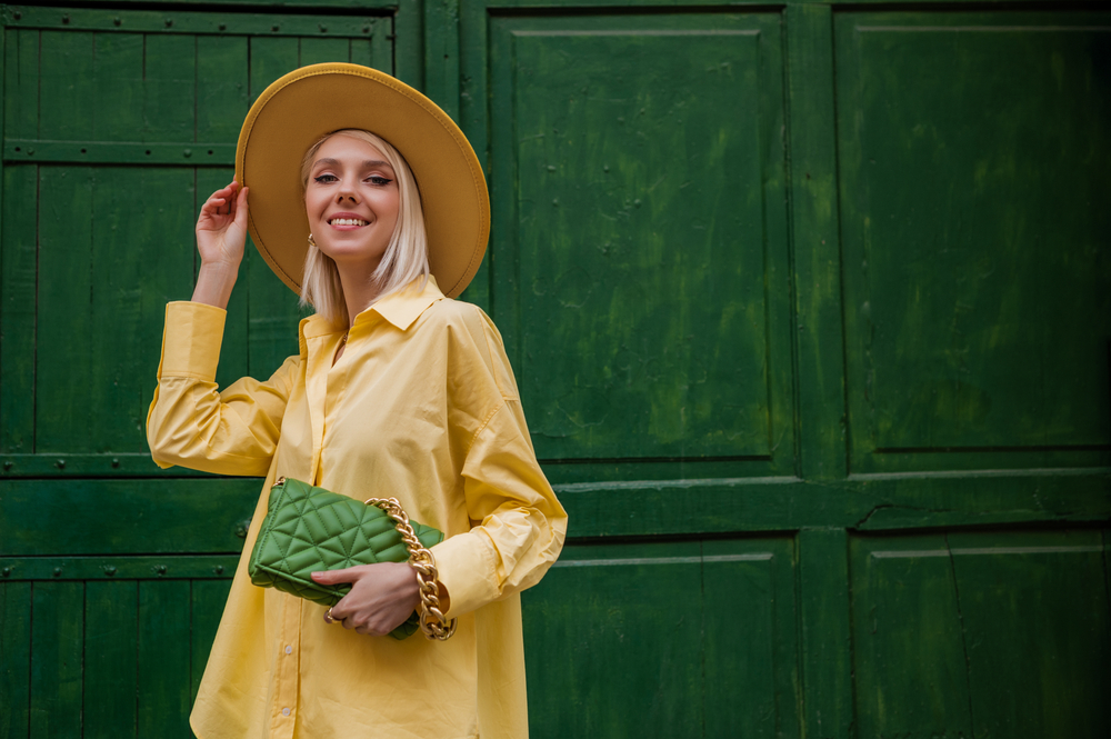 A woman tips her at as she stands in front of a green wall, her green purse matching the wall and contrasting her yellow button down: she knows What to wear in Italy in spring in color pallets and style!