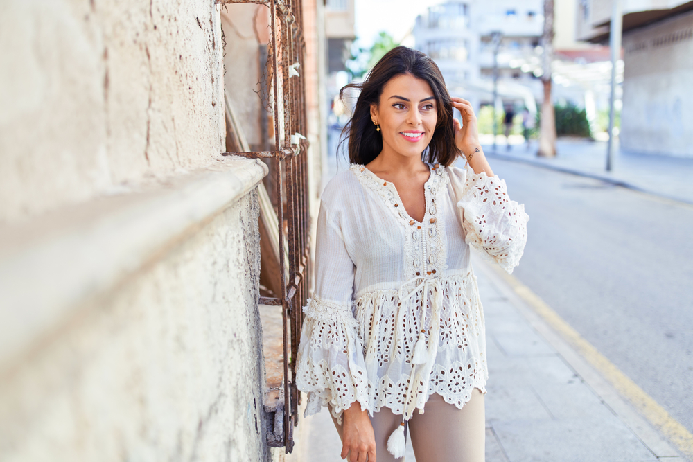 A woman smiles whole leaning against a wall of a building: she knows What to wear in Italy in spring in her flowy white blouse and chinos.