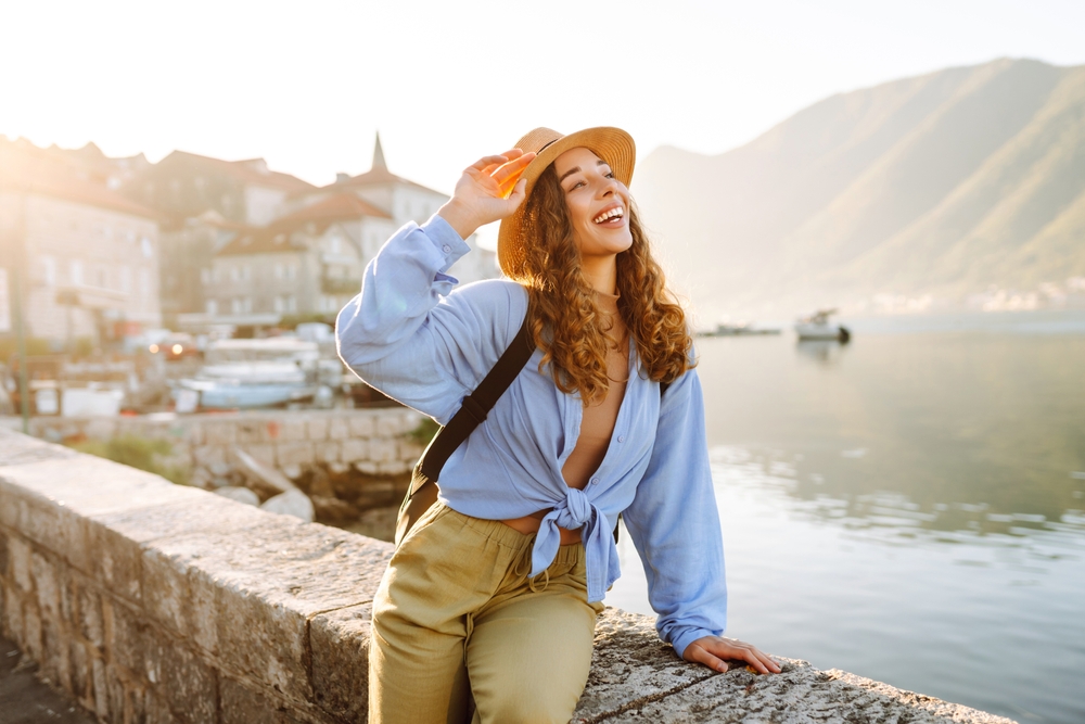 A woman smiles on the coast in her hat, joggers, and tied button down, she knows What to wear in Italy in spring!