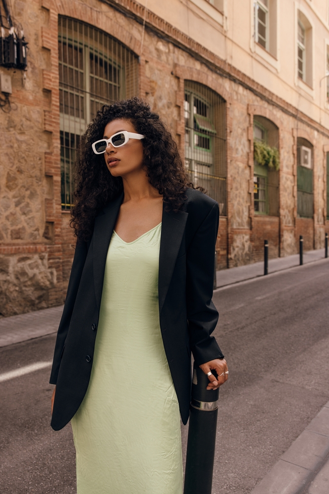 A woman walks down a street with stone buildings in a slip dress and a blazer with sunglasses.