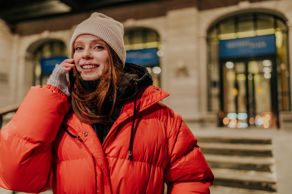 A woman smiles at the camera while on the phone, her parka, windbreaker and beanie keeping her warm.