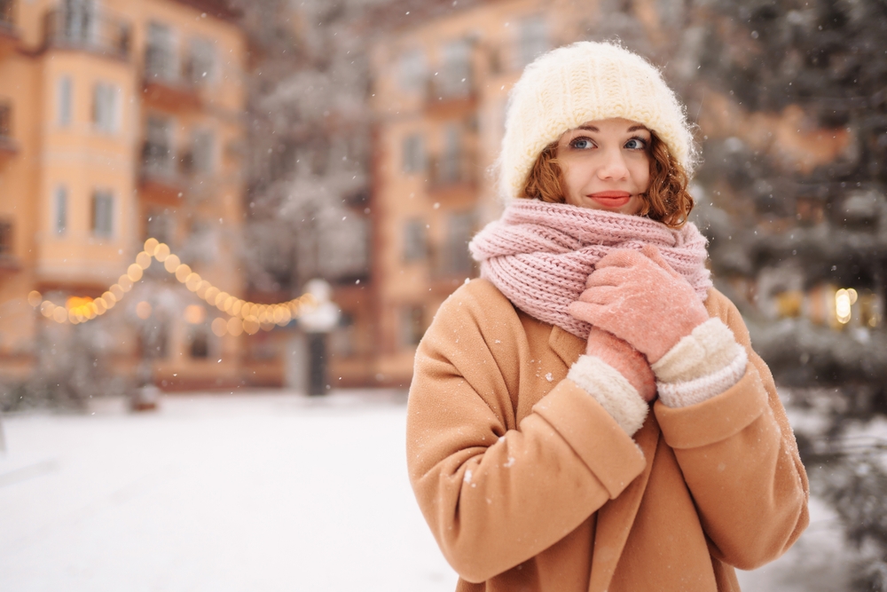 As it snows, a woman clutches her knit scarf to her chest, her wool coat, jacket, and gloves keeping her warm.