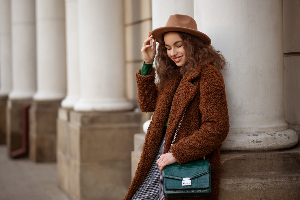 A young girl leans against a column and smiles while glancing down, her houndstooth pants and green top keeping her warm with her sherpa coat: she knows what to wear in Italy in winter.