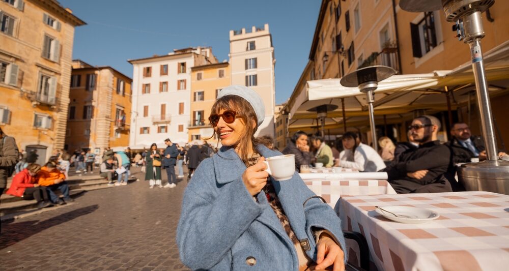 A woman leans back in her chair at an outdoor cafe, wearing a blue jacket and hat, knowing What to Wear in Italy in December to enjoy this outdoor seating.