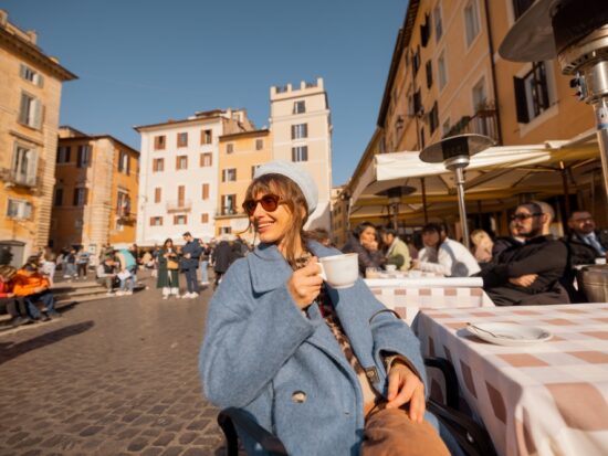 A woman leans back in her chair at an outdoor cafe, wearing a blue jacket and hat, knowing What to Wear in Italy in December to enjoy this outdoor seating.