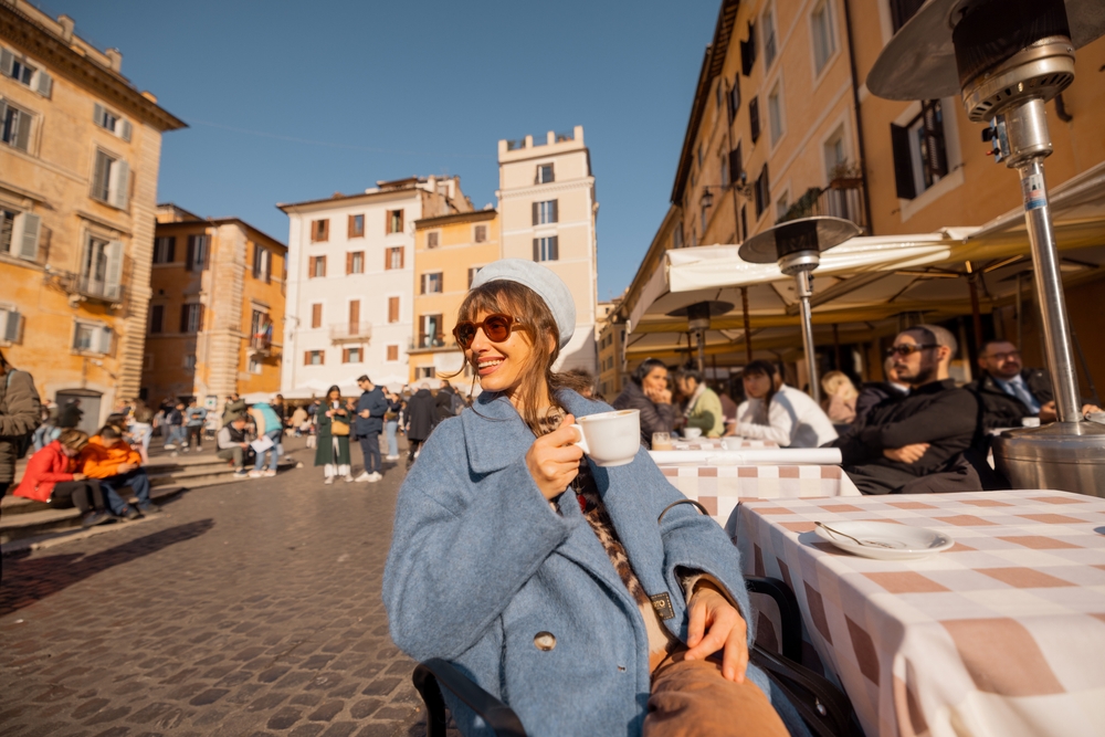 A woman leans back in her chair, knowing What to Wear in Italy in December, as she nurses coffee in an outdoor cafe in a blue wool coat.
