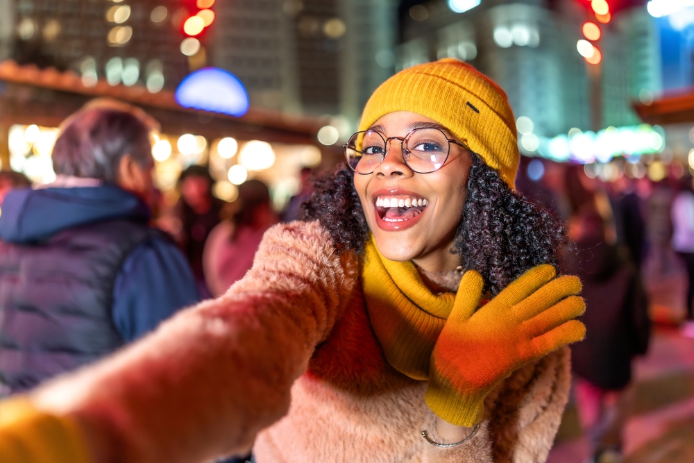 A selfie shows a girl waving at the camera in a fleece sweater, scarf, yellow gloves and beanie.