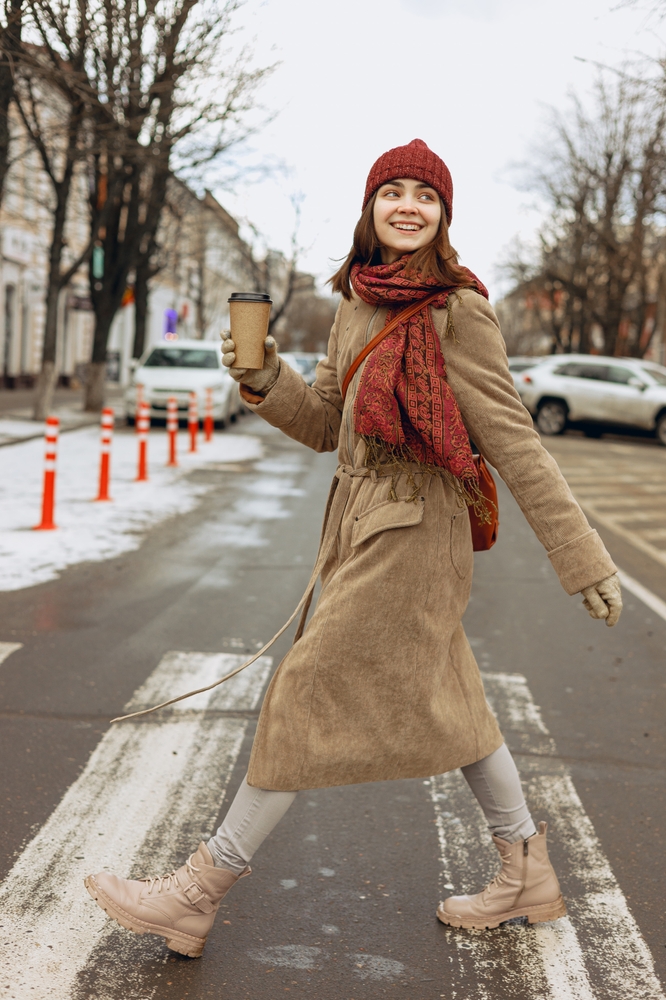 A woman crosses the street in a matching scarf and beanie with a long trench and coffee in her hand.