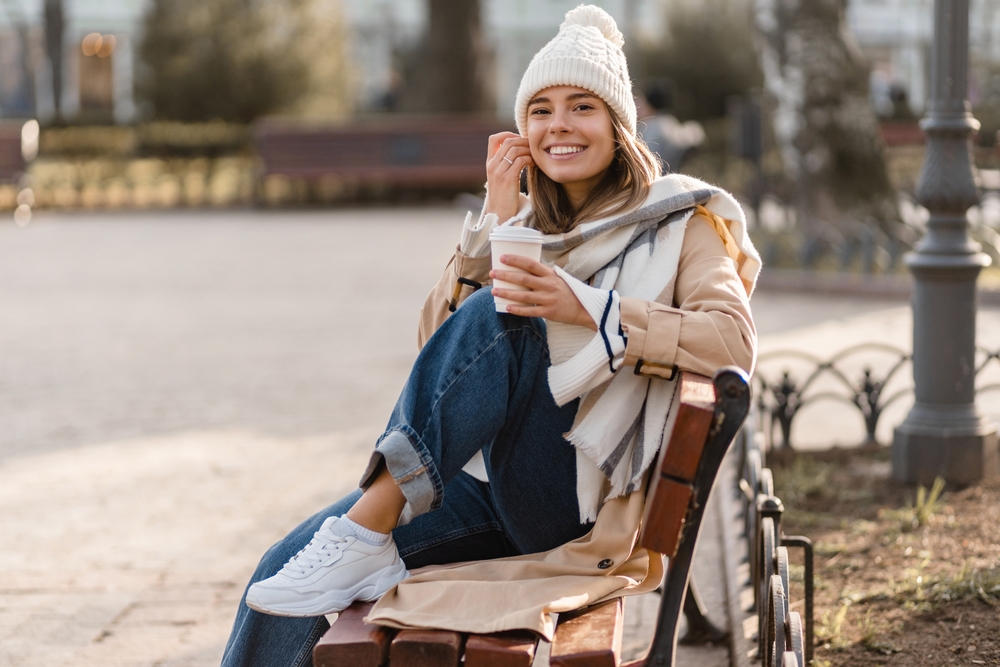 A woman knows What to Wear in Italy in December as she sits on a park bench in jeans, a beanie, and a scarf.