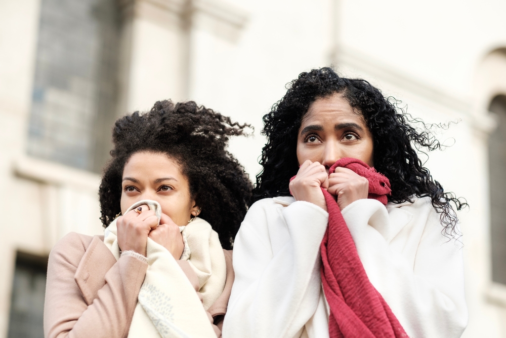 Two friends hold their scarves up to their noses as the wind rips through the city, their jackets not keeping them warm.