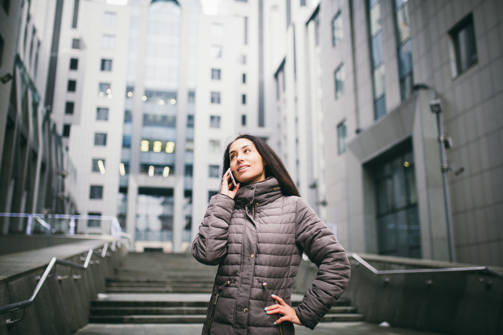 A business woman knows what to wear in New York City in December as she stands outside a city building while on her phone in a huge puffer jacket.