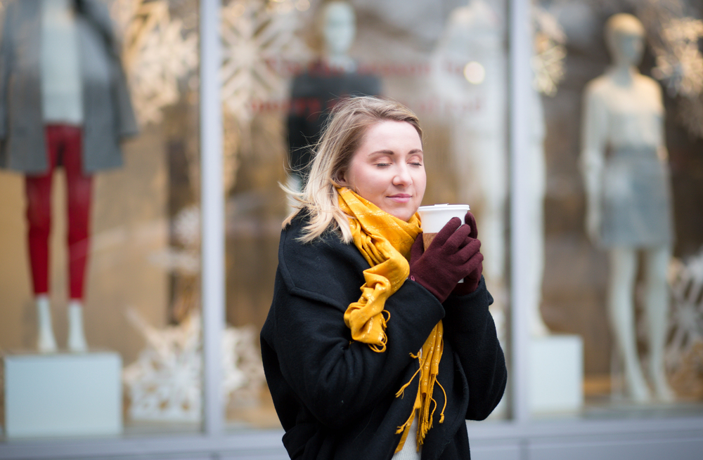 A woman holds tightly to a cup of coffee while outside: she knew what to wear in New York City in December with her wool parka, yellow scarf and thick gloves!