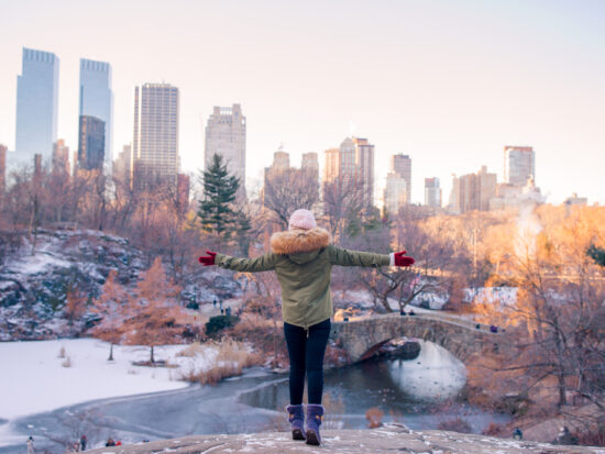 A woman knows what to wear in New York City in December as she looks over a frozen Central Park in a parka, thermal leggings, gloves and a beanie.