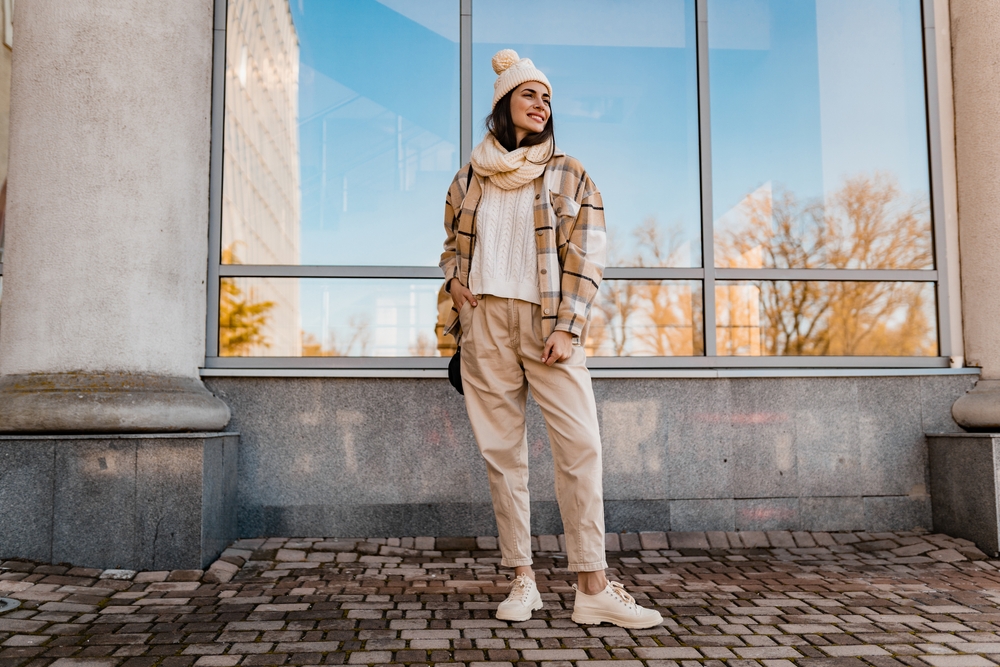 A woman poses in her trousers, sweater, scarf and beanie by a large window.