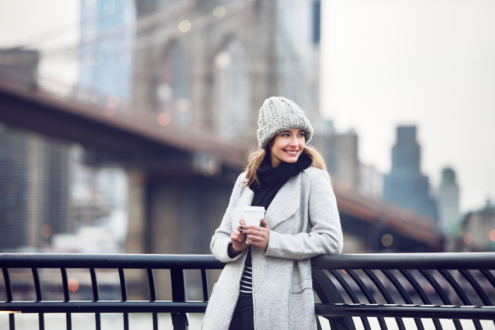A woman leans against a railing on the river in NYC, knowing what to wear in New York City in December in her wool parka, hat, scarf, and more.