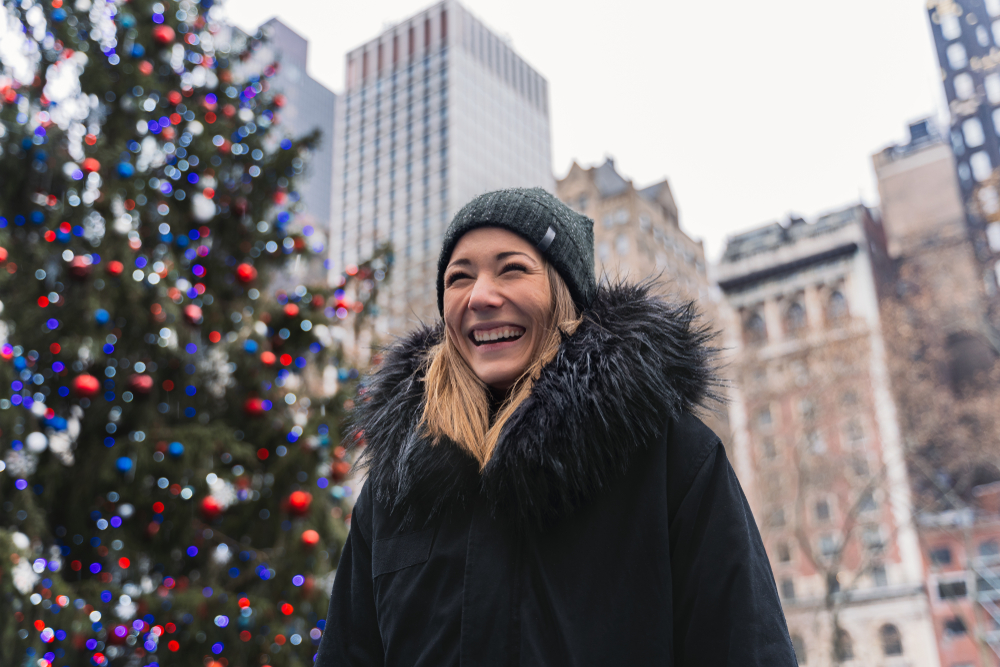 A girl knows what to wear in New York City in December as she laughs in her wool beanie and fur-lined parka in downtown by Christmas trees.