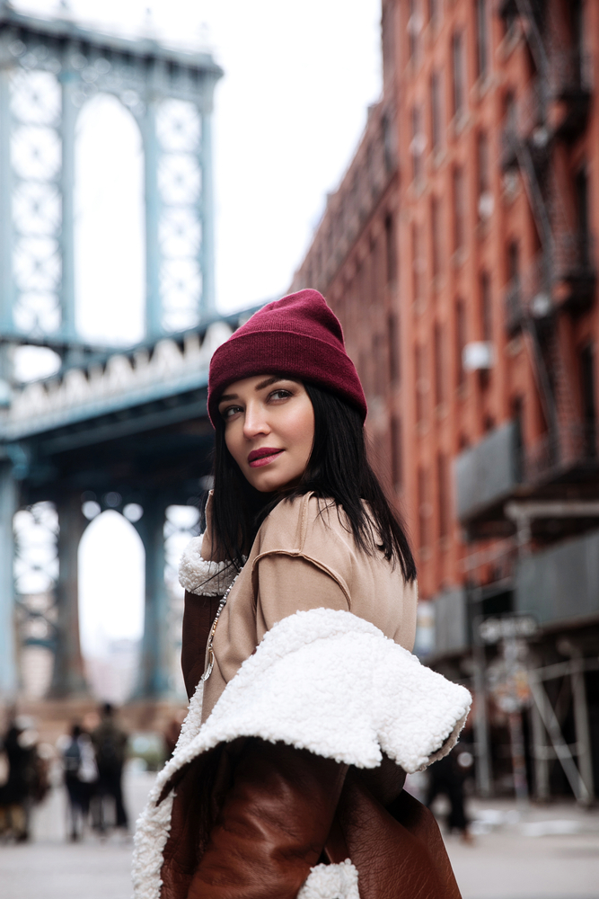 A fashionable girl knows what to wear in New York City in December, and poses over her shoulder in her read beanie, tan shirt, and fur-lined jacket.