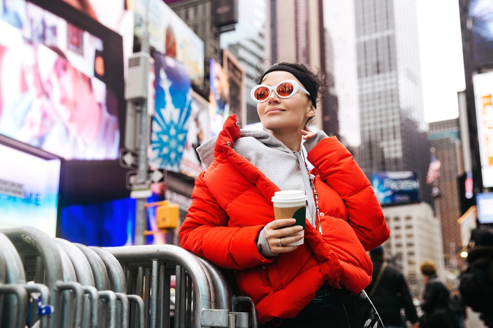 A woman in a hoodie and puffer jacket admires Times Square, knowing what to wear in New York City in December, as she nurses a coffee to keep warm.