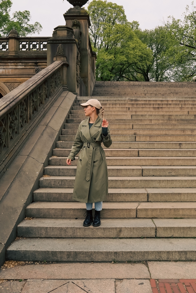 A woman descends a set of stairs in a long trench coat, jeans, a ball cap and boots to keep herself warm.