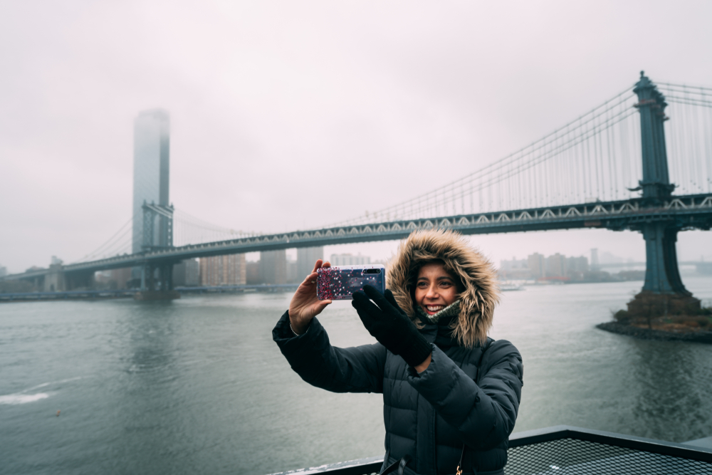 A woman in fur-lined parka takes a selfie with the Brooklyn bridge during a foggy, cold day.