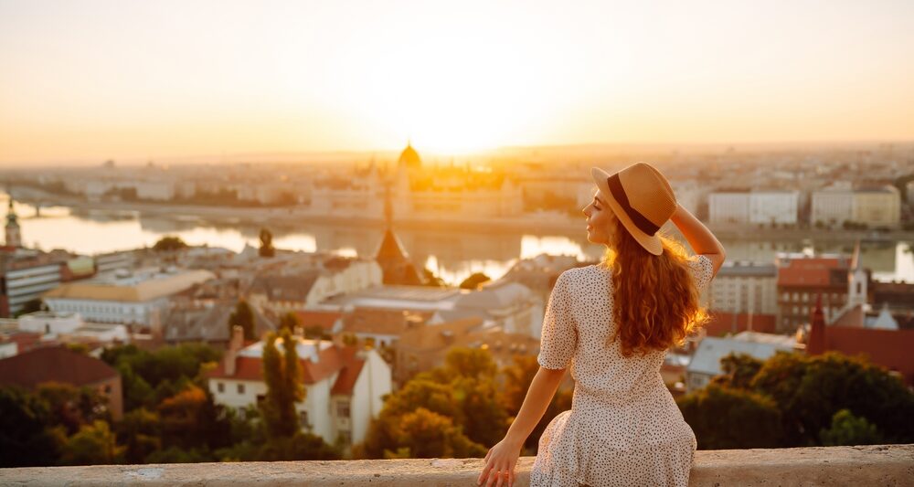 woman in a white dress sitting on a wall facing the city of budapest below. the woman has her hand on her tan hat and has her hair down. her hair is golden blonde. the sun is rising over the city of budapest and you can see buildings in the distance