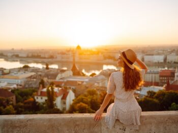 woman in a white dress sitting on a wall facing the city of budapest below. the woman has her hand on her tan hat and has her hair down. her hair is golden blonde. the sun is rising over the city of budapest and you can see buildings in the distance