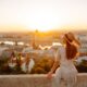 woman in a white dress sitting on a wall facing the city of budapest below. the woman has her hand on her tan hat and has her hair down. her hair is golden blonde. the sun is rising over the city of budapest and you can see buildings in the distance
