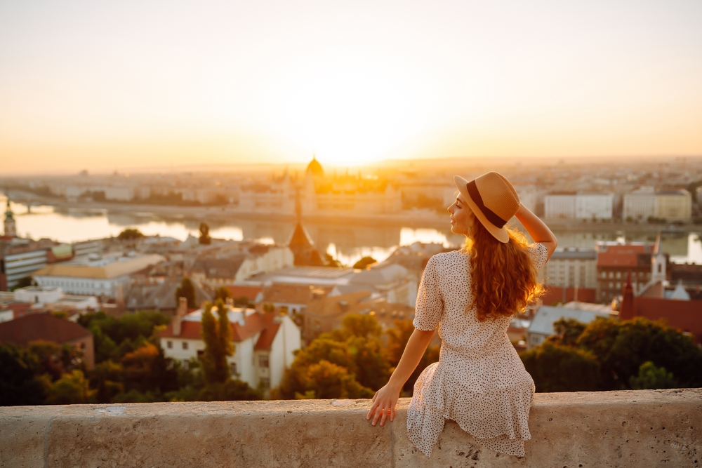 a woman is sitting on a stone ledge overlooking the danube river. she is wearing a sun dress and a sun hat, the sun is rising